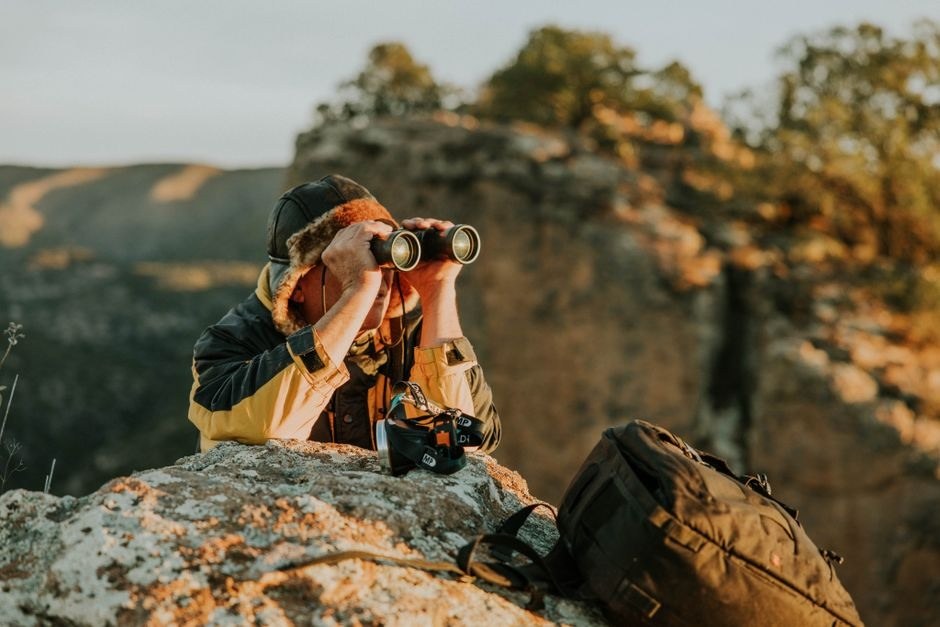 Photographer using binoculars from a rocky overlook at golden hour